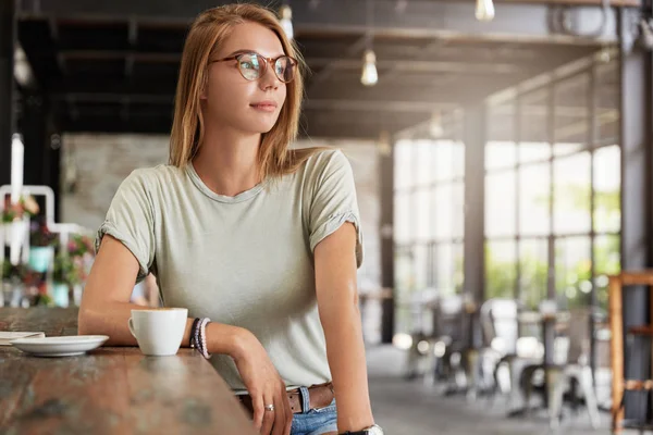 Indoor shot of attractive young female in eyewear sits at bar counter, enjoys tasty hot cappuccino or coffee, waits for friend, looks thoughtfully in window.