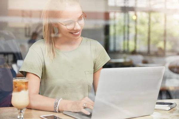 Beautiful female copywriter watches interesting video in websites, keyboards on laptop computer, sits at cozy cafeteria, enjoys tasty sweet cocktail.