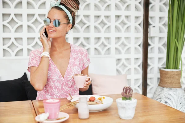 Stylish female wears shades, looks aside, holds cup of delicious espresso with foam, has pleasant conversation via modern cell phone.