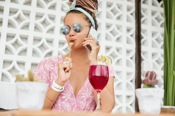 Fashionable young woman in trendy eyewear, licks teaspoon as eats something sweet and drinks cocktail or shake, speaks over modern smartphone, shares her impressions of spent holidays abroad