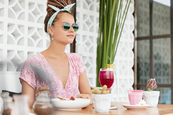 Female tourists with dreadlocks hairstyle, wears trendy round sunglasses, enjoys travelling in hot tropic country, has dinner in luxurious restaurant, eats exotic food and drinks fresh cool beverage