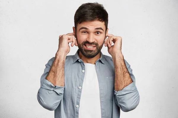 Young handsome man with stubble tries to ignore annoying noise, plugs ears, dressed casually, has displeased expression, isolated over white background. Male refuses to listen something unpleasant