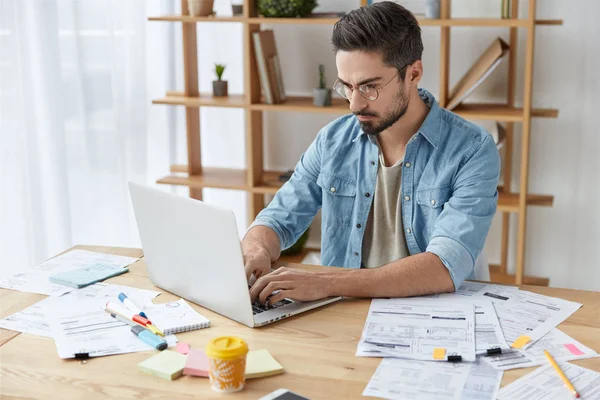 Indoor shot of concentrated brunet male surrounded with many documents in cozy cabinet, prepares financial project for business meeting. Talented writer keyboards new article on laptop computer