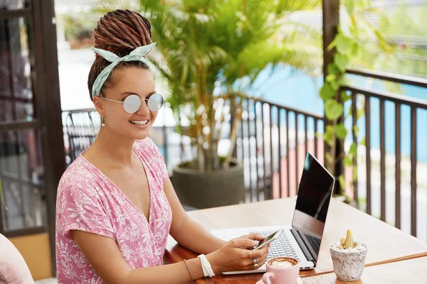 Stylish female with dreadlocks, wears stylish sunglasses, enjoys coffee at outdoor terrace cafe, rest in tropical country, receives text message on smartphone, uses portable laptop for communication