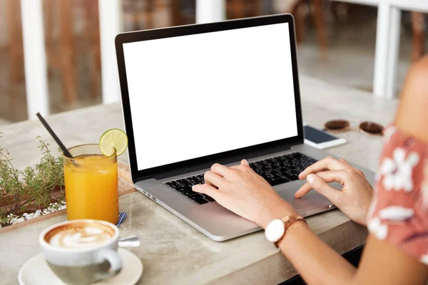 Cropped image of female employee types on keyboard of modern portable laptop computer with blank copy screen, sits at table with juice and cappuccino, works on financial report or checks email