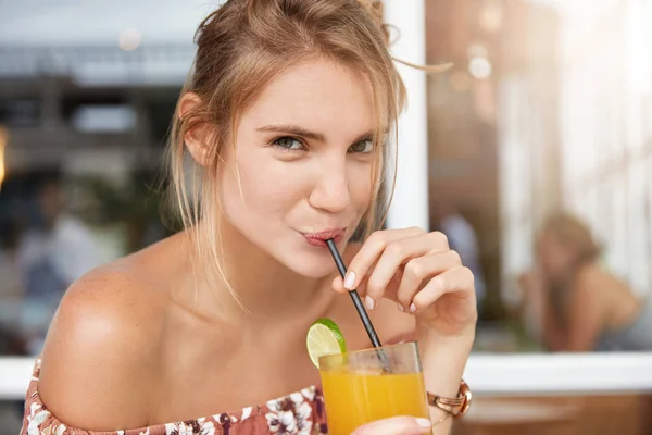 Adorable beautiful young female sips fresh orange juice from straw, wears fashionable clothing, spends summer holidays, sits at cafeteria interior, being thirsty.