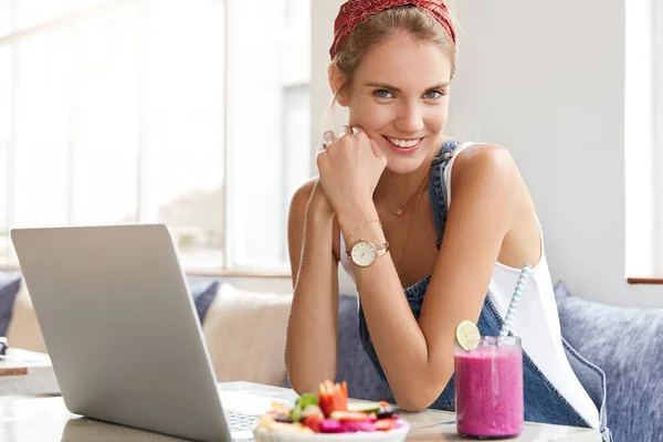Adorable female blogger works at laptop computer, thinks about creative ideas to develop her website, uses wireless internet, enjoys tasty shake and fruit salad.