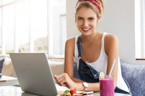 Fashionable young female wears headband and denim overalls, creates own network website on laptop computer, enjoys delicious cocktail, communicates online with customers, uses wireless internet