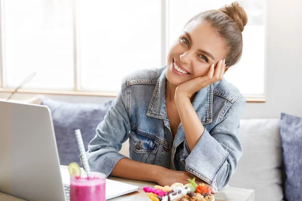 Smiling female student prepares for university quality control, being in good mood as enjoys tasty dessert. Beautiful blogger has free time at cozy cafe, uses laptop computer for publishing posts
