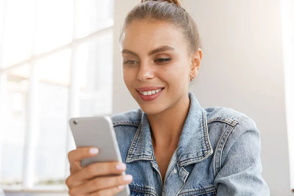 Pleased good looking female in stylish clothes, smiles happily, reads good news on mobile phone, being satisfied with high speed internet connection.