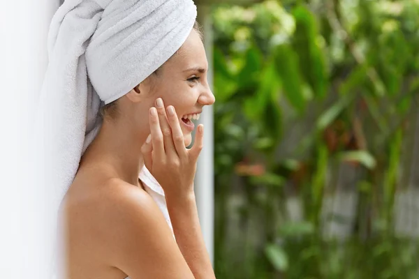 Sideways shot of happy pleased young female with white towel on head demonstrates her soft healthy skin after beauty treatments, poses against green tropical background.