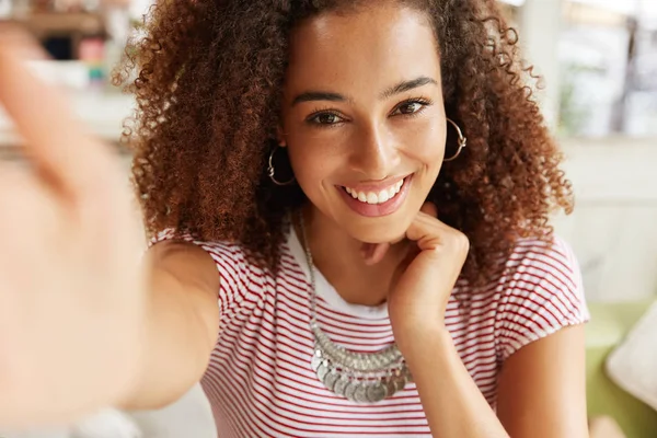 Adorable African American female model with frizzy dark hair, smiles at camera of her cell phone while makes selfie against cafe interior or makes video call, shares news with friends in social network