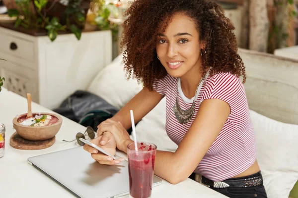 Mixed race black female model work freelance at cafe interior, surrounded with modern technologies and tasty drink. Attractive Afro American female browses information via smart phone online