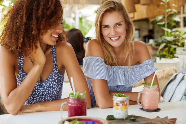 Horizontal shot of females best friends with broad smile sit together at cafe table, share impressions about summer holidays, drink tasty smoothie and eat exotic dish. People, lifestyle, friendship