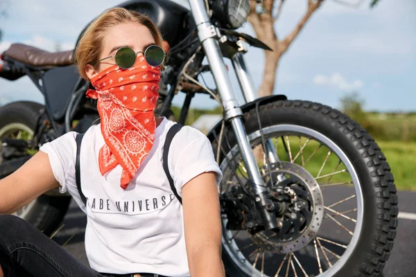 Young biker girl in white t shirt and sunglasses, sits at road near motorcycle, looks thoughtfully away, enjoys beautiful nature after covered long distance.