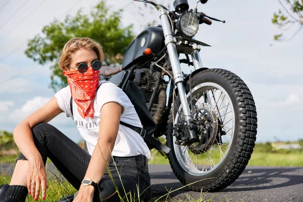 Outdoor shot of young female biker wears white t shirt, jeans and sunglasses, carries bag, feels free and relaxed as rides motorbike in unknown places.