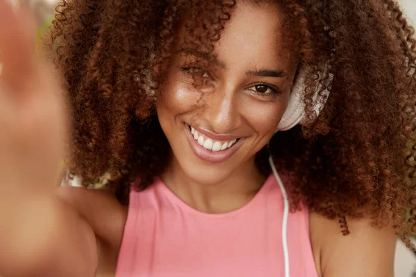 Portrait of curly beautiful African American woman in headphones, enjoys favorite music, makes photo of herself, has broad smile, dressed casually. Young dark skinned hipster girl poses for selfie