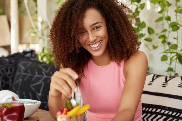 Curly woman with cheerful expression, eats delicious dessert, being in good mood, spends spare time at cozy coffee shop, enjoys tasty fruit salad. Attractive female rests after excursion alone