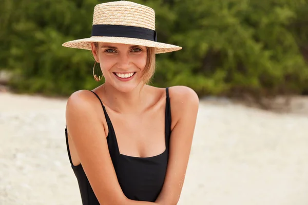 Bright picture of woman looks happily directly into camera, demonstrates her perfect slim body, wears bikini and hat, bathes in sun during day time at summer, relaxes outdoor in tropical beach