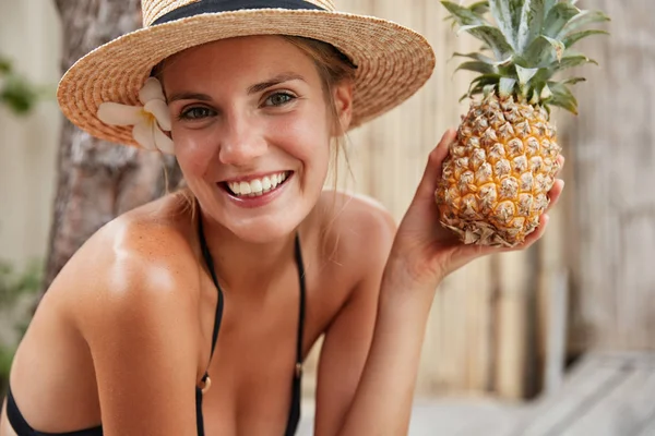 Gorgeous happy female in bikini and summer hat relaxes on spa in tropical hotel, holds pineapple, prepares for party with friends. People, healthy eating, fruit diet and recreation concept.