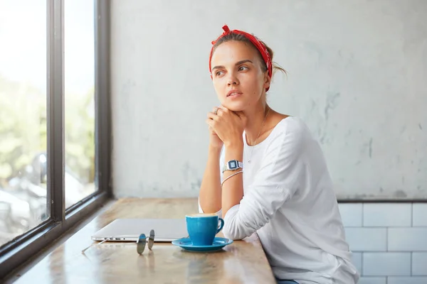 Thoughtful cute female blogger dressed in casual clothing, sits at coffee shop, contemplates about something as looks into window, uses laptop computer, drinks hot beverage, has break after work
