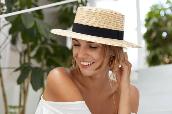 Pretty glad female wears straw hat, shows her tanned bare shoulder, smiles with happy expression as looks joyfully aside, spends summer vacations abroad. Lovely young woman enjoys good resort alone