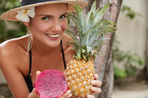 Positive carefree female wears straw hat, keeps exotic fruits in hands, rests at tropical country, has tanned skin, broad smile. Beautiful woman with pineapple and dragon fruit. Tourism, recreation