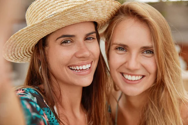 Close up shot of adorable females have friendly relationships, make selfie, have broad smiles, stand close to each other.