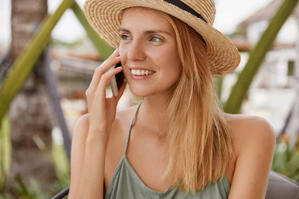 Portrait of happy blonde female wears straw hat and happy smile, looks positively away, has pleasant talk with boyfriend via cell phone, shares impressions about being on beach resort. People and talk