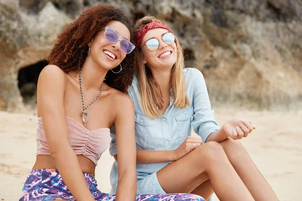 Happy friends enjoys recreation time at desert with cheerful expressions, wear sunglasses, sit against cliff background. Young lovely feminists enjoys togetherness in calm resort place