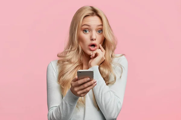 Beautiful female with blonde hair feels surprised as checks her email box on cell phone, wonders big discounts in web store, isolated over pink background. Emotions and unexpectedness concept