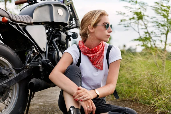 Pensive female biker deep in thoughts wears fashionable clothing, looks away with thoughtful expression, sits on ground near motorbike, covers long destination.