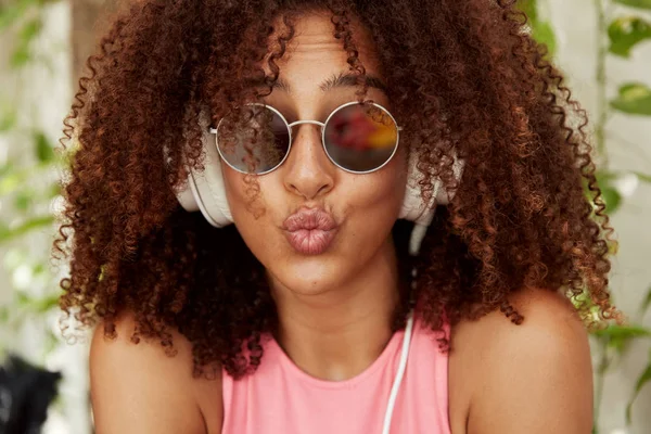 Funny African American woman with rounded lips, makes grimace, has curly bushy hair healthy dark skin, wears sunglasses, listens audio track in modern white headphones.