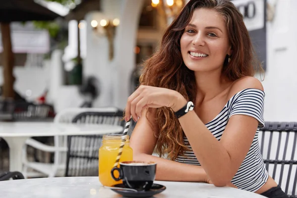 Satisfied young woman with pleasant appearance recreates at coffee shop, happy to spend summer holidays with boyfriend in exotic country, drinks fresh orange juice and cappuccino.