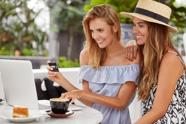 Portrait of happy best female friends pay with credit card, use banking application on modern laptop computer, make orders, drink aromatic coffee with piece of cake.