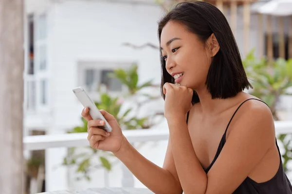 Photo of glad Asian woman watches funny video on social networks via cell phone, sits in outdoor cafeteria, texts message, uses free wifi, updates information.