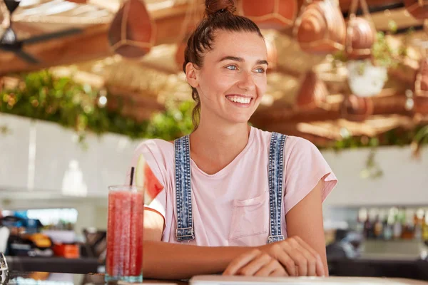 Portrait of adorable blue eyed female model with hair knot wears overalls, enjoys drinking fruit smoothie, looks happily somewhere while sits in cozy restaurant.
