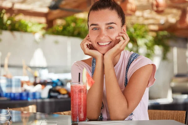 Glad young female with satisfied expression, has attractive look, keeps hands under chin, being relaxed as spend free time in cozy restaurant, enjoys fresh drink, has summer vacation in tropics