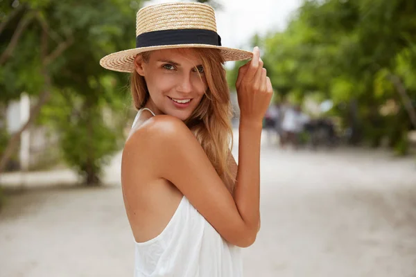 Outdoor shot of pleasant looking female with tanned healthy skin, dressed in white dress and summer hat, poses in park with confident satisfied expression, likes recreation.