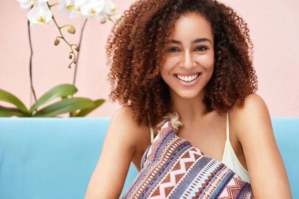 Glad dark skinned curly female with broad shining smile, embraces cushion while sits on comfortable couch against pink wall. Pretty adorable woman enjoys spare time at home.