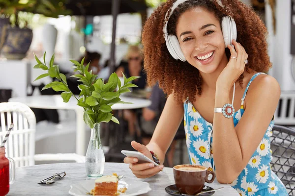 Dark skinned African American woman enjoys perfect sound of favorite song in headphones, connected to cell phone and chooses audio in playlist, drinks coffee in restaurant and eats dessert.