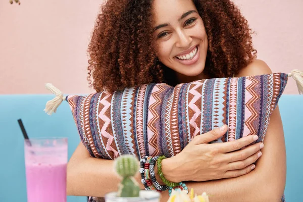 Joyful dark skinned female with positive expression spends good time in cafeteria together with boyfriend or close friend, embraces soft cushion, drinks cocktail, has rest in cozy cafeteria.