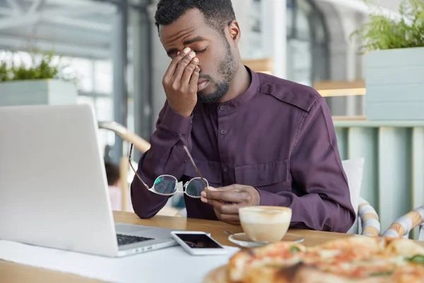 Overworked professional male economist, holds spectacles, being tired to work at laptop computer much hours, has headache after tired working day, sits at cafe with coffee, uses modern technologies