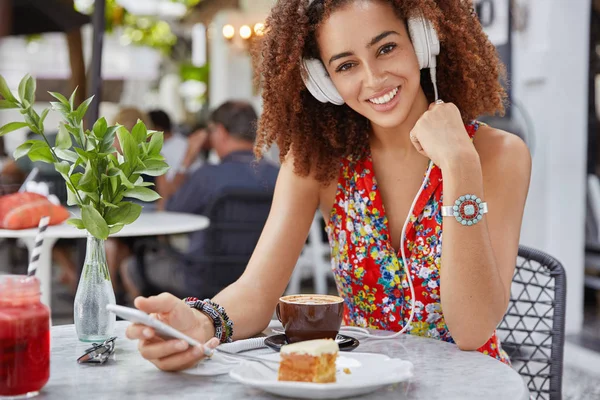Outdoor shot of delighted happy dark skinned young female wears red blouse, listens audio song in headphones, connected to wireless internet, enjoys tasty cake and aromatic coffee.