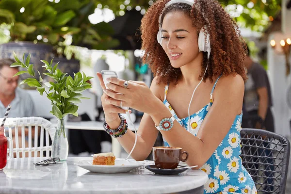 Glad concentrated African American female enjoys favorite song from playlist, connected to smart phone and headphones, sits in outdoor coffee shop, enjoys tasty dessert, updates application
