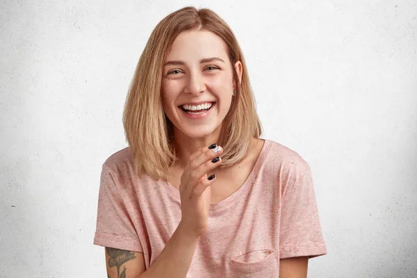 Horizontal shot of pleasant looking female laughs at good joke, being in high spirit, has broad smile, dressed casually, has manicure, isolated over white concrete background.