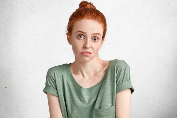 Headshot of discontent uncertain doubtful female with red hair tied in bun, hesitates about something, being displeased with everything poses against white background, demonstrates uncertainty