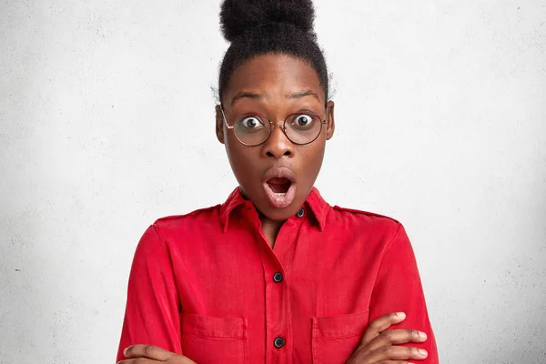 Beautiful emotional dark skinned businesswoman wears red formal blouse and spectacles, stares at camera with shocked expression, has deadline to prepare business project.