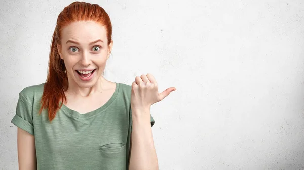 Excited woman looks with surprised amazed expression and bated breath, has ginger pony tail, indicates at blank copy space on white concrete background for your advertisement or promotional text