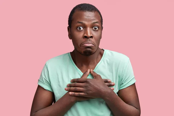 Puzzled dark skinned hesitant African American male keeps hands on chest, looks with widely opened eyes and curves lips, expresses doubt and disbelief, isolated over pink studio background.